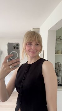 Černý top bez rukávů s detailním knoflíkem, hladká látka a boční rozparek. Osoba drží telefon v průhledném obalu, v pozadí jsou police.