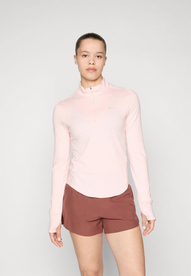 Lys rosa langermet topp med kvart glidelås og Nike-logo, sammen med brune treningsshorts, som viser glatt, elastisk stoff.