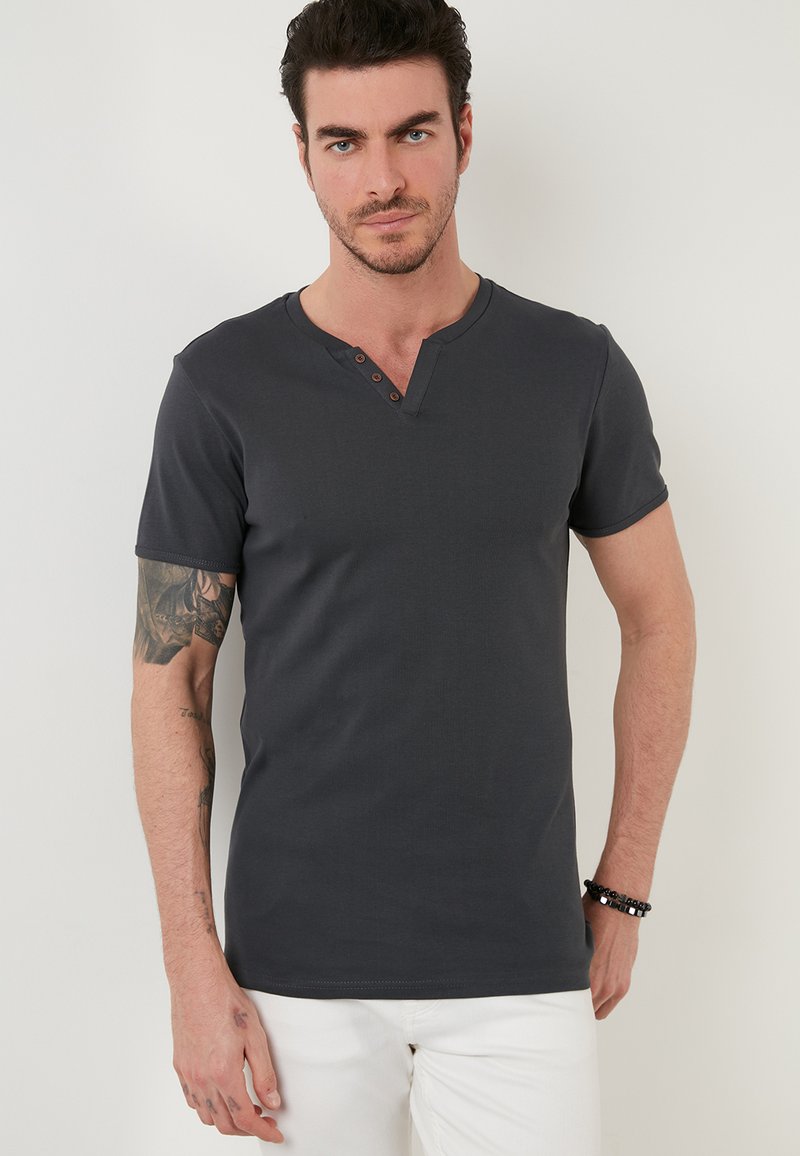 Tricou gri cu mânecă scurtă, cu decolteu în V și trei nasturi maro la guler. Textură netedă, design ajustat. Purtați cu pantaloni albi.