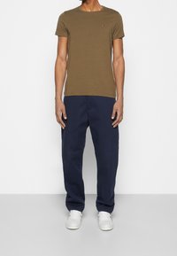 Oliivinvihreä lyhythihainen t-paita pyöreällä pääntiellä ja pienellä logodetaljilla, yhdistettynä löysälahkeisiin tummansinisiin housuihin ja valkoisiin lenkkareihin.