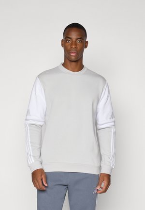 Sweatshirt gris clair avec des manches blanches, présentant trois bandes verticales sur chaque bras. Tissu doux et poignets côtelés, design à col rond.