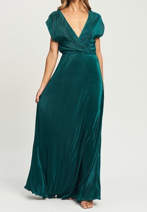 Robe maxi plissée couleur sarcelle avec un décolleté en V profond, manches courtes bouffantes et taille cintrée, portée par une personne debout tenant la jupe d'une main.