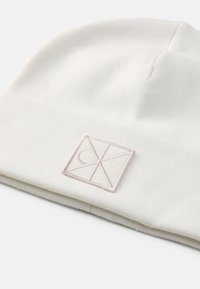 Bílá pletená �čepice s ohrnutým lemem. Na přední straně je jemně vyšitý geometrický logo v bledě růžové barvě. Měkká textura, jednoduchý design.