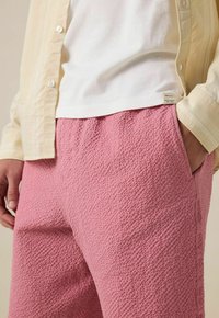 Pantalons roses texturés avec un motif de surface prononcé, associés à une chemise blanche et une chemise à boutons jaune clair.