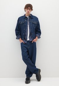 Giacca di jeans con tasche anteriori, abbinata a una camicia a quadri e a una T-shirt bianca. Pantaloni di jeans a vestibilità ampia e scarpe nere con suole testurizzate.