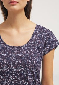 Mujer que lleva una blusa de manga corta azul marino con un patrón de puntos dispersos en blanco y rojo y un escote redondo.