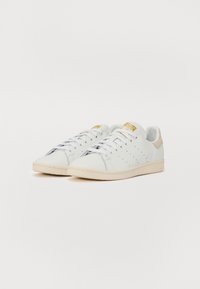 Zapatillas de piel blanca con detalles perforados, cordones planos, suela de goma color crema y un acento en el talón en tono marrón. Branding dorado en la lengua.