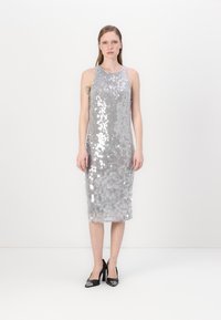 Vestido de lantejoulas prateadas com um design justo, corte sem mangas, comprimento até aos joelhos e textura leve; combinado com sapatos de salto pretos.