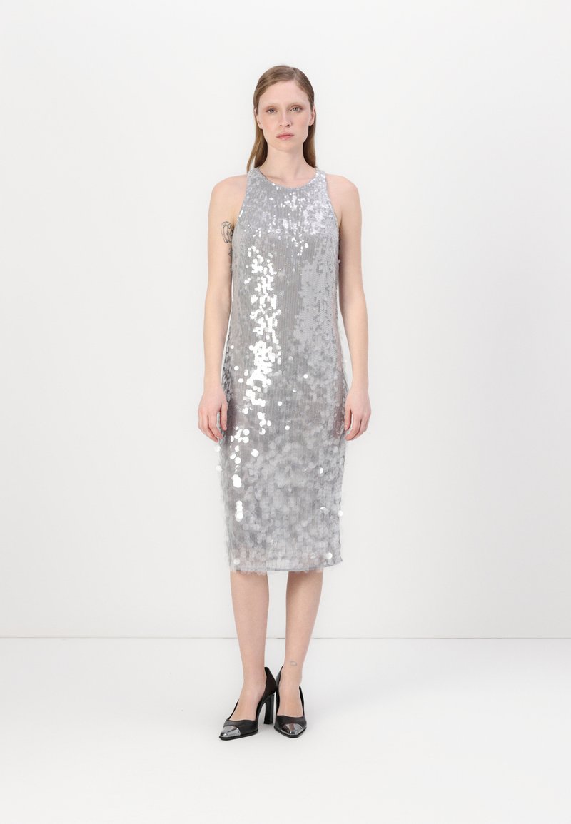 Vestido de lantejoulas prateadas com um design justo, corte sem mangas, comprimento até aos joelhos e textura leve; combinado com sapatos de salto pretos.