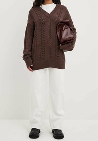 Brauner gestrickter Pullover mit V-Ausschnitt, der über Zopfmuster verfügt. Kombiniert mit weißen Hosen und einer burgunderfarbenen Tasche. Schwarzes Schuhwerk.
