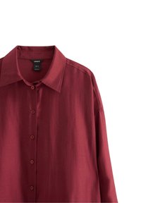 Burgundská košile s dlouhým rukávem a límečkem, se šesti knoflíky a viditelnou černou etiketou uvnitř výstřihu.