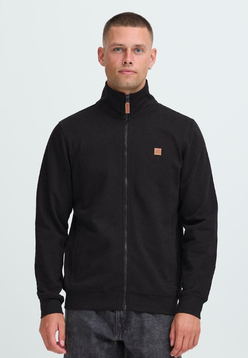 Mann med kort hår som har p�å seg en svart jakke med høy krage og liten brun logo, står mot en enkel lys bakgrunn.
