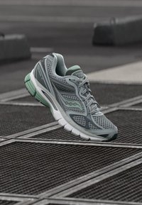 Grauer Laufschuh aus Netzstoff mit einem mintgrünen Akzent. Verfügt über eine gedämpfte Sohle und ein leichtes Design, schwebt in der Luft vor einem strukturierten Hintergrund.