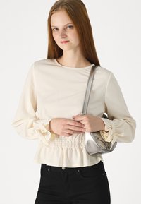 Jeune femme aux longs cheveux roux portant un chemisier crème à poignets évasés et un pantalon noir, tenant un sac à épaule en argent.