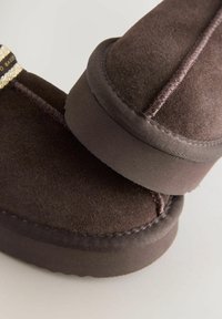 Pruunist sametist saapad ümarate kujunditega, millel on kontrastne õmblemine ja tekstureeritud kummitald. Pehme tekstuur ja minimalistlik disain.