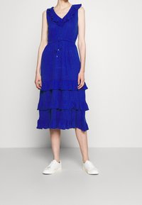 Blå ärmlös klänning med volangdetaljer, knapprad fram, justerbart midjeband och lager på lager-skirt. Bärs med vita sneakers.