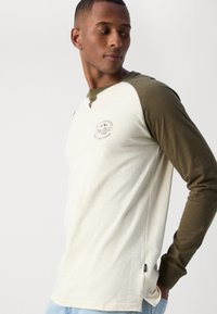 Pikade värvitud rinhaavaga T-särk kreemikas keha ja oliivroheliste varrukatega, millel on rinnal väike trükitud logo. Pehme kangas ja külgedel lõhikuga.