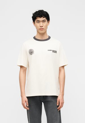 Νέος άνδρας με κοντά σγουρά μαύρα μαλλιά, φοράει λευκή μπλούζα με μαύρα λογότυπα κειμένου και σκουρόχρωμο γκρι τζιν, σε απλό φόντο.