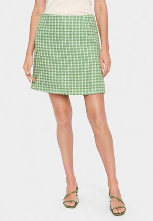 Grüner Tweedrock mit figurbetonter Form, strukturierter Oberfläche und weißem Karomuster. Über dem Knie geschnitten, kombiniert mit grünen Riemchensandalen.