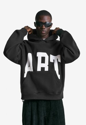 Zwarte hoodie met grote witte "ART" opdruk. Voorzien van een voorzak en een trekkoord in de capuchon. Draagt met donkere, gestructureerde broek.