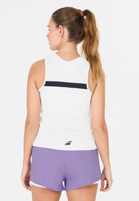 Donna con coda di cavallo marrone che indossa un top bianco senza maniche con striscia nera e pantaloni sportivi viola, rivolta di spalle contro uno sfondo bianco.