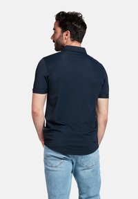 Polo bleu marine à manches courtes, en tissu lisse, avec un détail de empiècement au dos. Associé à un jean bleu clair, se tenant les mains dans les poches.