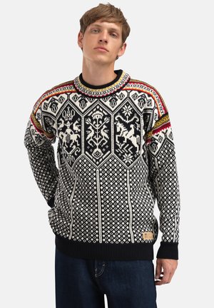 Ung mann iført en strikkegenser i svart, hvitt, rødt og gult med geometriske og dyremønstre, stående med én hånd på hoften.