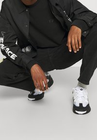 Svart jacka med vita grafiska tryck, svart skjorta och svarta jeans. Skor: kraftiga svarta och vita sneakers med texturerad yta.