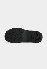Semelle de chaussure en caoutchouc noir avec des motifs de bande de roulement profonds, triangulaires et rectangulaires pour l’adhérence, montrée sur un fond gris clair uni.