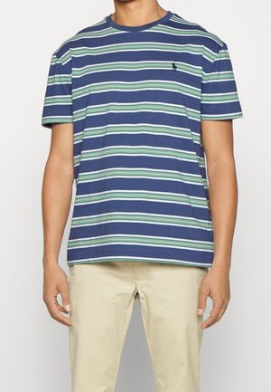 Mand iført en kortærmet T-shirt med blå, grønne og hvide vandrette striber og beige bukser, stående foran en ensfarvet baggrund.