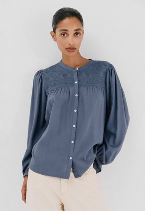 Blauwe blouse met knopen en puffed sleeves, met textuur borduursel op de borst en gladde stof. Bevat witte knopen aan de voorkant.