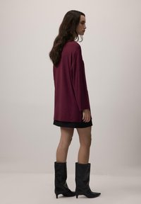 Sieviete ar gariem tumšiem matiem stāv sānis, ģērbusies brīvā maroon krāsas augšpusē, melnā svārku un ceļgala augstuma melnās zābakos uz vienkārša fona.