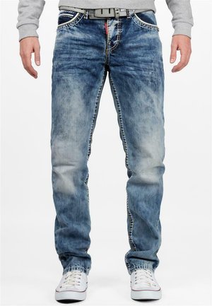 Jean en denim bleu délavé avec une coupe droite, texture effilochée, coutures contrastantes et fermeture éclair, associés à des baskets blanches.