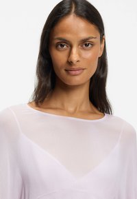 Lyserød, gennemsigtig bluse med lange ærmer, en subtil struktureret mønster og en rund halsudskæring. Har en trekantet detalje foran.