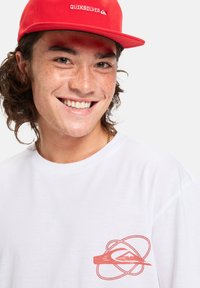 Baltas marškinėlis su koralinio atspalvio Quiksilver logotipu kairėje krūtinės pusėje ir raudona kepurė. Audinys atrodo lygus, o kepurė turi lygią kraštą.