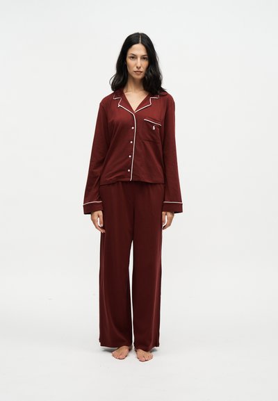 Femme debout pieds nus, portant un haut de pyjama rouge foncé à manches longues boutonné et un pantalon de pyjama ample assorti, sur un fond blanc uni.