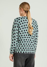 Kobieta z długim blond kucykiem, nosząca zielony sweter w czarno-białe okrągłe wzory oraz ciemnoszare dżinsy, zwrócona plecami.