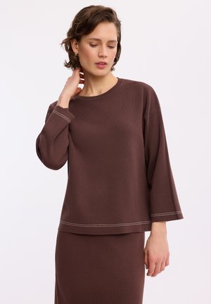 Kobieta ubrana w brązową, luźną bluzkę z długim rękawem z białymi detalami szwów i dopasowaną spódnicę, pozująca z ręką przy szyi.