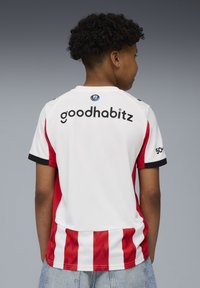 Ung person med kort lockigt hår som b�är en vit och röd sporttröja med "goodhabitz"-logga på ryggen.