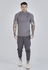 Pelēks sporta krekls ar īsām piedurknēm, ar ribotu tekstūru un logotipu uz krūtīm, apvienots ar pelēkiem jogginga biksēm un baltām kedām.