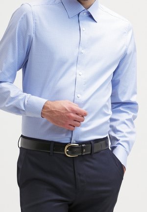 Mann, der ein hellblau kariertes Hemd mit Knöpfen trägt, in dunkle Hosen mit schwarzem Gürtel und goldener Schnalle gesteckt, Hand in der Tasche.