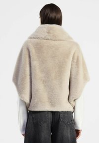 Donna che indossa una giacca in pelliccia sintetica beige a maniche corte sopra una maglietta bianca a maniche lunghe e jeans in denim grigio scuro, vista da dietro.