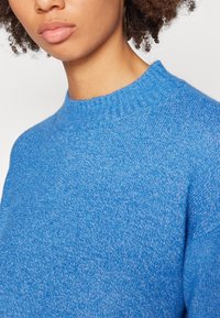 Blauer Strickpullover aus weichem, texturiertem Stoff. Mit einem gerippten Kragen und einer lockeren Passform, die ein leicht marmoriertes Muster aufweist.