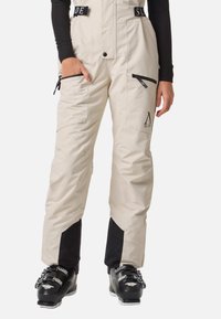 Pantalons de ski beige avec accents noirs, dotés de plusieurs poches zippées, de bretelles ajustables et d'une coupe fuselée ; matériau résistant à l'eau.