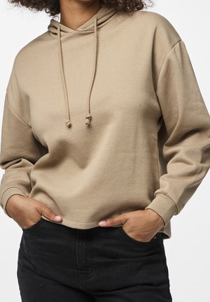 Personne portant un sweat à capuche beige avec des cordons de serrage et un pantalon noir, les mains glissées dans les poches avant sur un fond uni.