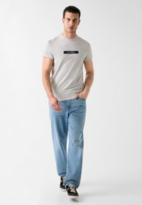 T-shirt gris clair avec un logo "GUESS" noir, assortie à un jean droit bleu clair et des baskets noires avec des lacets blancs.