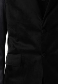 Blazer de terciopelo negro con una textura suave, dos botones y un bolsillo de ojal, que presenta un diseño elegante sin patrones visibles.
