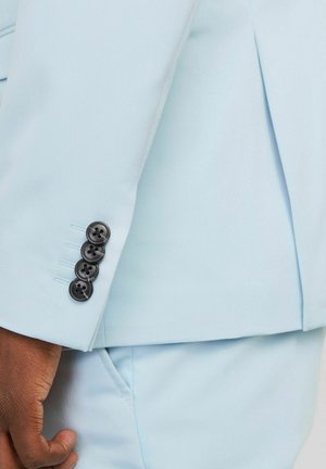 Main dans la poche d'un costume bleu clair, gros plan sur la manche de la veste avec quatre boutons foncés.