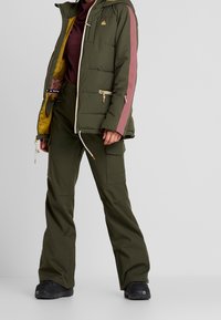 En person som bär en grön puffy-jacka med vinröda ränder, ihop med gröna byxor och svarta stövlar, står mot en neutral bakgrund.