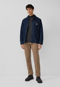 Giovane uomo in piedi che indossa una giacca blu scura, un maglione grigio a collo alto, pantaloni beige e stivali neri su uno sfondo chiaro e uniforme.